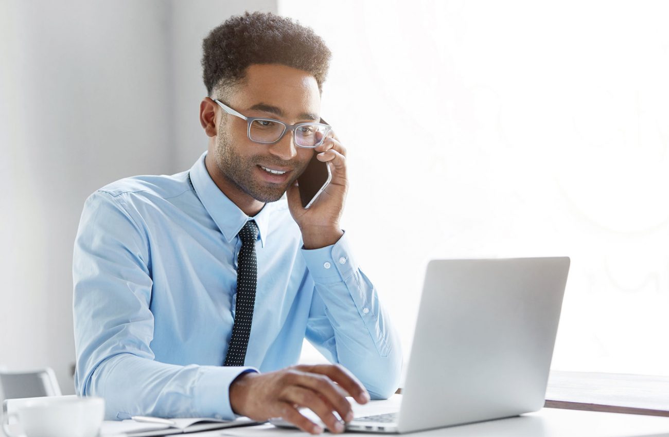 Businesspeople, modern technology and communication concept. Good looking unshaven young African American male employee working on generic laptop computer and having phone conversation in office